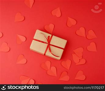 box wrapped in brown kraft paper and tied with a red thin silk ribbon, cut out of paper hearts, red background, gift for Valentine&rsquo;s Day, birthday, flat lay