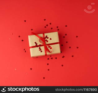 box wrapped in brown kraft paper and tied with red thin silk ribbon on a red background , decorative shiny hearts are scattered nearby, holiday is Valentine&rsquo;s Day