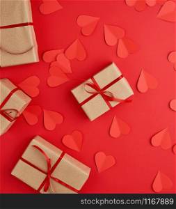 boxes wrapped in brown kraft paper and tied with red thin silk ribbon on a red background, decorative hearts are scattered nearby, holiday is Valentine&rsquo;s Day