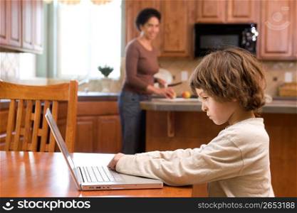 Boy (4-5) using laptop while mother cutting fruits in kitchen