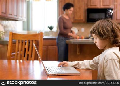 Boy (4-5) using laptop while mother cutting fruits in kitchen