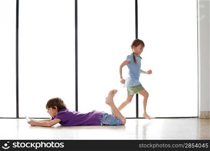 Boy (7-9) lying on floor playing handheld game girl (5-6) running