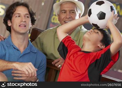 boy balancing a soccer all on his head with his father and grandfather looking at him