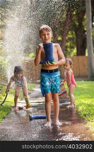 Boy carrying welly on sidewalk, girls playing with water hose in background