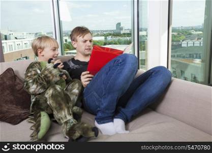 Boy dressed in dinosaur costume sitting with father reading story book on sofa bed at home