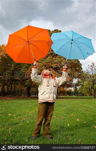 Boy in autumn park. Holds over head two colour umbrellas under cloudy sky.