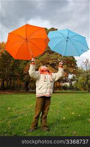 Boy in autumn park. Holds over head two colour umbrellas under cloudy sky.
