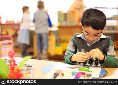 Boy in glasses moulds from plasticine on table in kindergarten