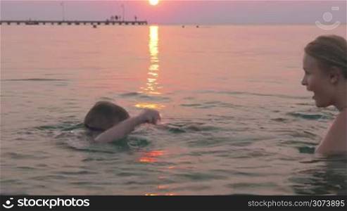 Boy in goggles learning to swim with his mother against of beautiful sunset