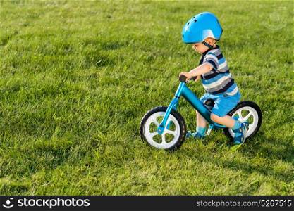 Boy in helmet riding a blue balance bike (run bike). Happy child learning to keep balance on a training bicycle in the garden. Healthy preschool children summer activity. Kid playing outside. First day on the bike.