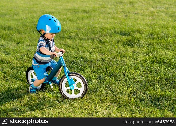 Boy in helmet riding a blue balance bike (run bike). Happy child learning to keep balance on a training bicycle in the garden. Healthy preschool children summer activity. Kid playing outside. First day on the bike.