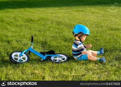 Boy in helmet sitting on grass with blue balance bike (run bike). Healthy preschool children summer activity. Kid playing outside. First day on the bike.