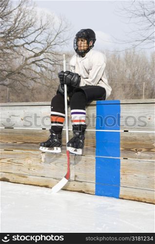 Boy in ice hockey uniform holding hockey stick sitting on sidelines.