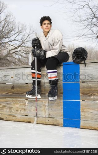 Boy in ice hockey uniform holding hockey stick sitting on sidelines.
