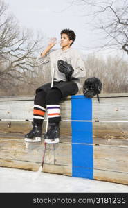 Boy in ice hockey uniform holding hockey stick sitting on sidelines drinking water.