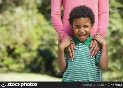 Boy in park making funny expression with mother&acute;s hands on shoulders.