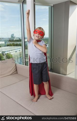 Boy in super hero costume standing with arm raised on sofa bed at home