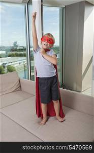 Boy in super hero costume standing with arm raised on sofa bed at home