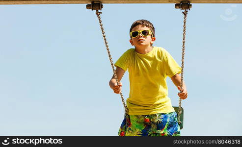 Boy playing swinging by swing-set.. Rest and relax for children. Little boy in sunglasses resting swinging outdoor. Adorable child having fun playing in playground.