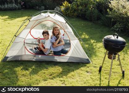 boy playing ukulele sitting back back his sister tent near barbecue grill