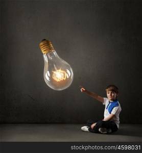 Boy pointing idea. Cute school boy sitting on floor and touching glass light bulb with finger