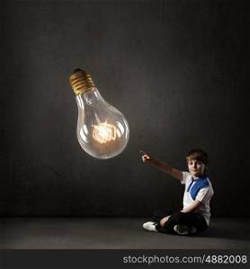Boy pointing idea. Cute school boy sitting on floor and touching glass light bulb with finger
