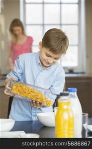 Boy pouring corn flakes in bowl with mother standing in background