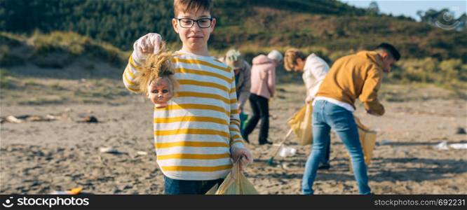 Boy showing doll head with disgust face while volunteer group cleans the beach. Boy showing doll head while cleaning the beach