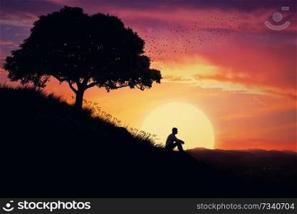 Boy sit alone on a hill in the center of nature, over a sunset background. Standing away from the crowd, waiting for the healing power of the nature.