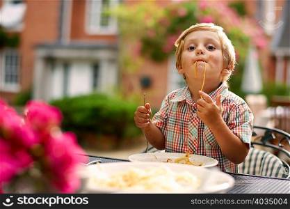 Boy sitting at garden table sucking up spaghetti looking at camera