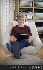 Boy Sitting In Chair In Bedroom At Home Wewaring Headphones And Playing On Digital Tablet
