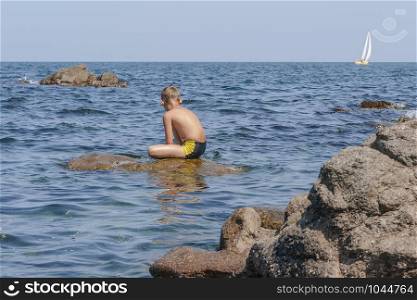 Boy sitting on a big stone in the sea, jutting out of the water