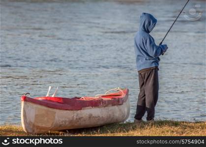 Boy, standing next to canoe on the banks of a river, with fishing rod in hand, busy fishing.