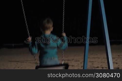 Boy swinging alone on the beach playground. Evening scene with dark sea in front of a kid