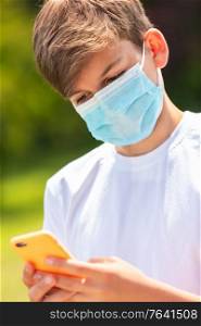 Boy teenager teen male child wearing a face mask outside during the Coronavirus COVID-19 virus pandemic while using his mobile cell phone for social media