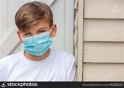 Boy teenager teen male child wearing a face mask outside leaning against the side of a house during the Coronavirus COVID-19 virus pandemic