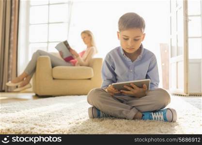 Boy using digital tablet on floor with mother reading magazine in background