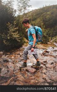 Boy with backpack going across a mountain stream during hiking in mountains