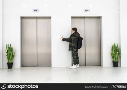 Boy with casual clothes and white sneakers call the elevator. White contemporary building interior. Flowers in pots and white wall. Metallic elevator doors.