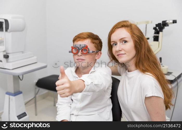 boy with optometrist trial frame showing thumb up gesture sitting with young female ophthalmologist clinic