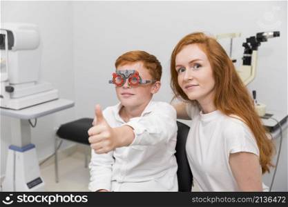 boy with optometrist trial frame showing thumb up gesture sitting with young female ophthalmologist clinic