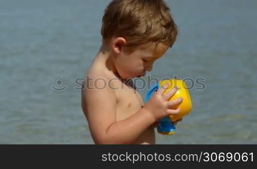 Boy with toy waterpot on the beach, then focus on mother having tea and enjoying sunny day, her son in background