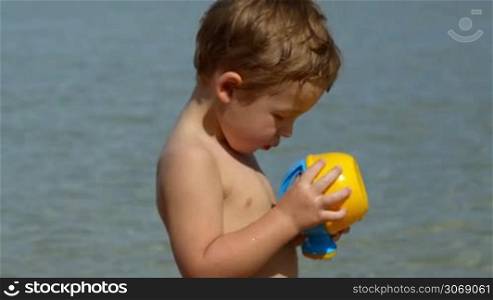 Boy with toy waterpot on the beach, then focus on mother having tea and enjoying sunny day, her son in background