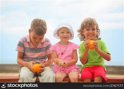 boys and girl with oranges and one liittle girl with panama hat is sitting on wooden bench. boy wearing stripped t-shirt looking at orange. focus on face of girl with orange.