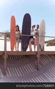 Boys and girls teen surfers with surfboards standing on a wooden pathway looking to the sea waves at the beach.