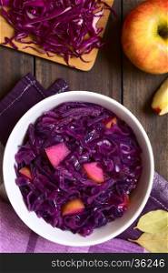 Braised red cabbage with apple in bowl surrounded by ingredients, photographed overhead on dark wood with natural light (Selective Focus, Focus on the top of the dish)