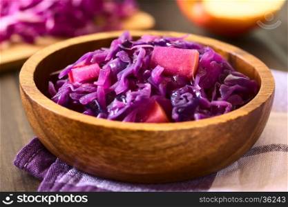 Braised red cabbage with apple in wooden bowl, with ingredients in the back, photographed with natural light (Selective Focus, Focus in the middle of the dish)