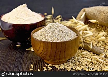 Bran flakes oat and oat flour in two wooden bowl, a bag of grain oats, oat ears against the dark wooden board