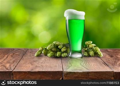 Branch of natural organic hop with glass of fresh cold green beer on a wooden table against natural blurred background, copy space. Happy St.Patrick &rsquo;s Day concept.. Still life with glass of fresh green beer and hops.