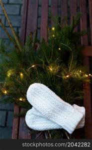 Branches of a coniferous tree decorated with a garland lie on a bench along with knitted white mittens. Place for an inscription. Branches of a coniferous tree decorated with a garland lie on a bench along with knitted white mittens. Place for an inscription.